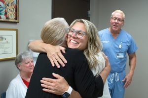 Dr. Marnie Kaplan smiles as she hugs a woman patient, who is seen from behind. Dr. Chance Kaplan, standing on the right, and a woman, seated on the left, look on. Dr. Marnie Kaplan has long hair and is wearing glasses and a top. The woman patient is wearing a sweater. Dr. Chance Kaplan is wearing scrubs and has a name badge on a lanyard attached to his pocket.