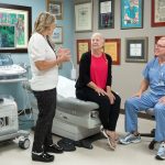 Dr. Marnie Kaplan, left, talks to a woman patient who is sitting on an exam table. Dr. Chance Kaplan, right, looks on. Dr. Marnie Kaplan is wearing a white top and black pants. The woman patient has a braid in her hair and is wearing a top and sweater. Dr. Chance Kaplan is wearing scrubs and has a name badge on a lanyard attached to his pocket.