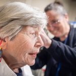 A woman is shown in profile as Dr. Jackie Price, an audiologist at Penn State Health Otolaryngology – Head and Neck Surgery, fits her with a hearing aid. A cord is hanging from the woman’s ear, and small ribbons hang from the hearing aid. Price, who is seen out of focus, is wearing scrubs, a lanyard and glasses and is smiling.
