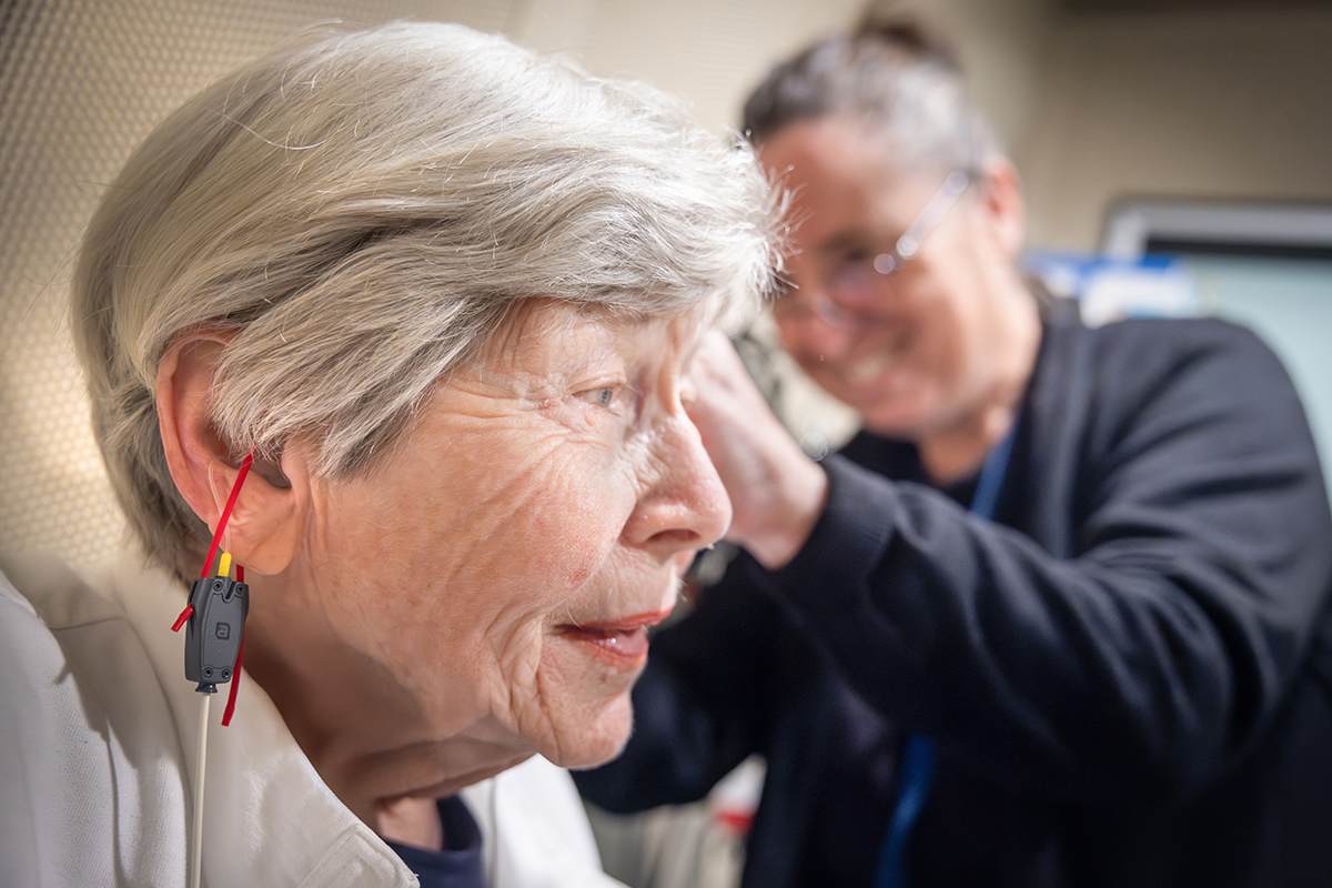 A woman is shown in profile as Dr. Jackie Price, an audiologist at Penn State Health Otolaryngology – Head and Neck Surgery, fits her with a hearing aid. A cord is hanging from the woman’s ear, and small ribbons hang from the hearing aid. Price, who is seen out of focus, is wearing scrubs, a lanyard and glasses and is smiling.