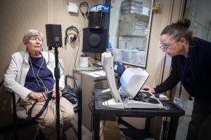 A woman sits in a chair on the left with cords hanging from hearing aids in her ears. Dr. Jackie Price, right, leans over a computer. Behind them are speakers, headphones and other audiology equipment.