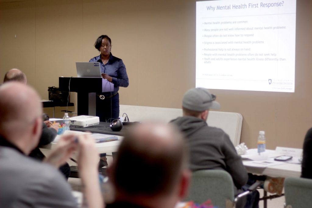 Ruth Moore of Pennsylvania Psychiatric Institute stands at a lectern teaching a class to a group of men. A PowerPoint slide on the wall is titled “Why Mental Health First Response?”