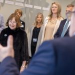 A group of people form a semi-circle around a woman who smiles as she looks at a man in a suit standing near the camera.