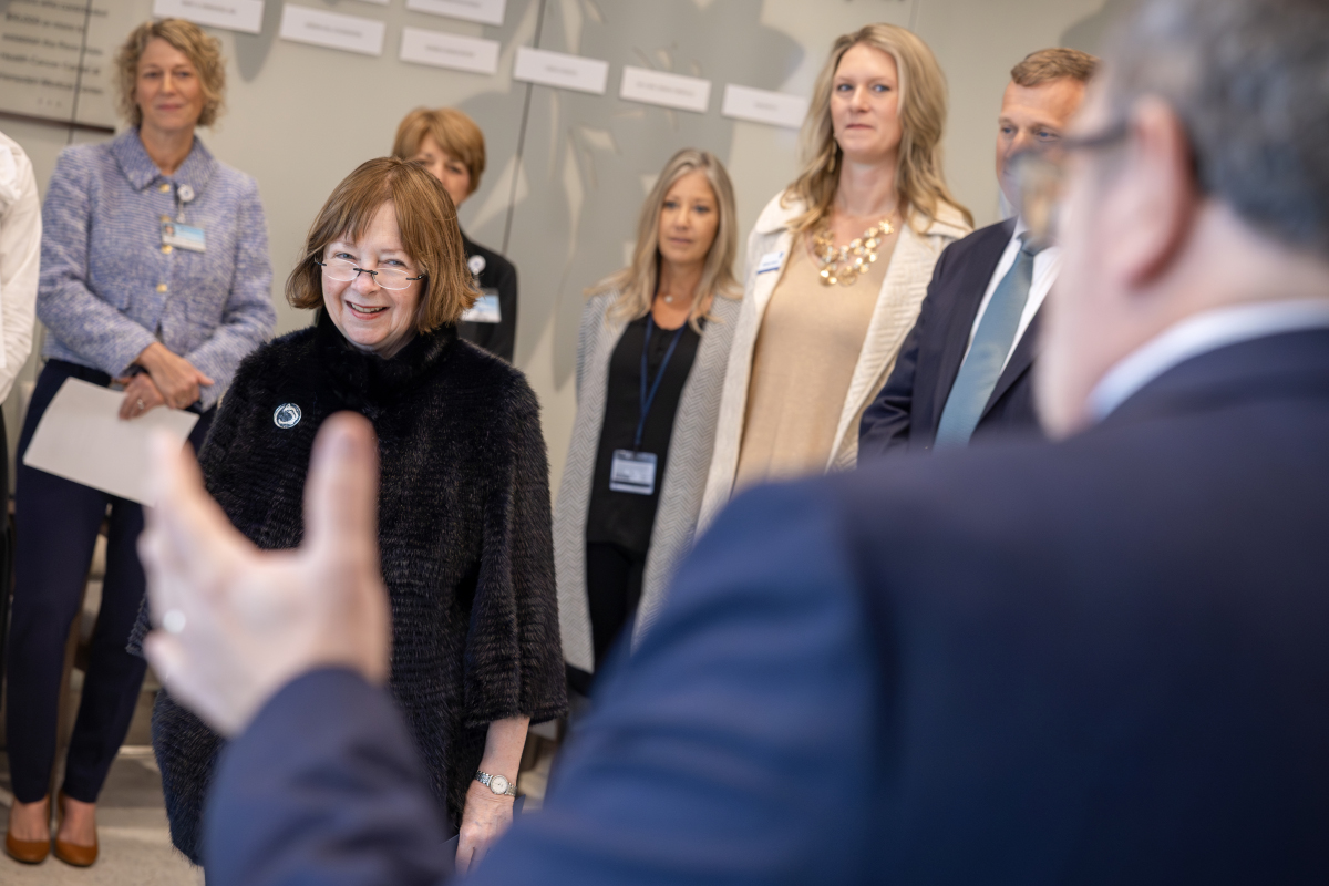 A group of people form a semi-circle around a woman who smiles as she looks at a man in a suit standing near the camera.