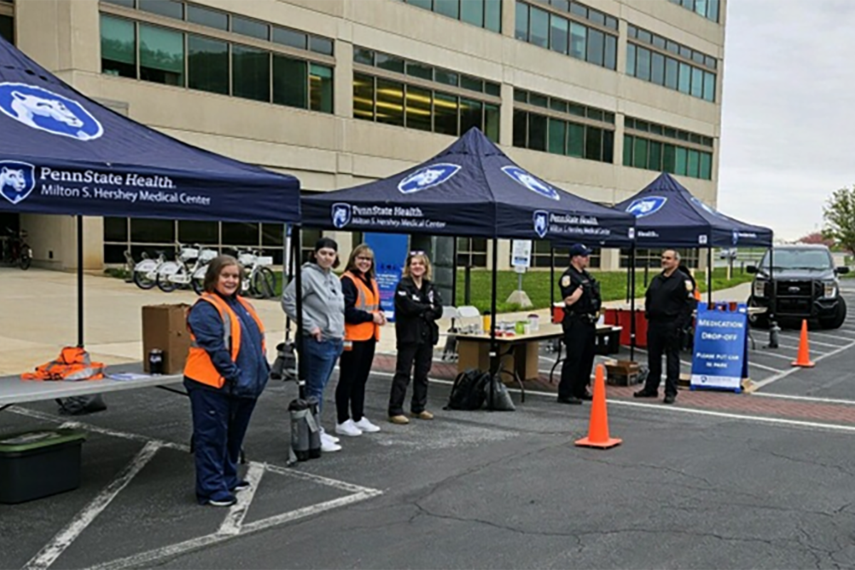 Three canopies branded Penn State Health Milton S. Hershey Medical Center a set up along the road in front of the medical center. Three hospital staff and three law enforcement officers stand under and around the canopies, facing the road.