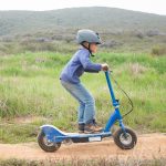 A young boy wearing a helmet rides an electric scooter on a dirt path.