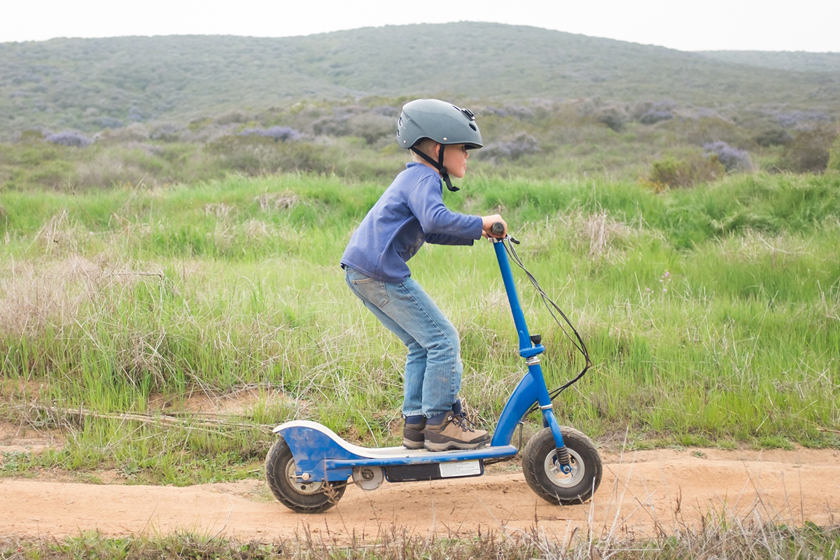 A young boy wearing a helmet rides an electric scooter on a dirt path.