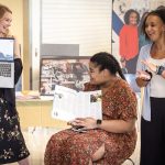 Three women smile and interact during a presentation. One woman stands on the left holding a laptop showing a person on a video call. Another woman sits in the center holding open a magazine article. The third woman stands on the right, clapping and smiling. Posters and display boards are visible in the background.