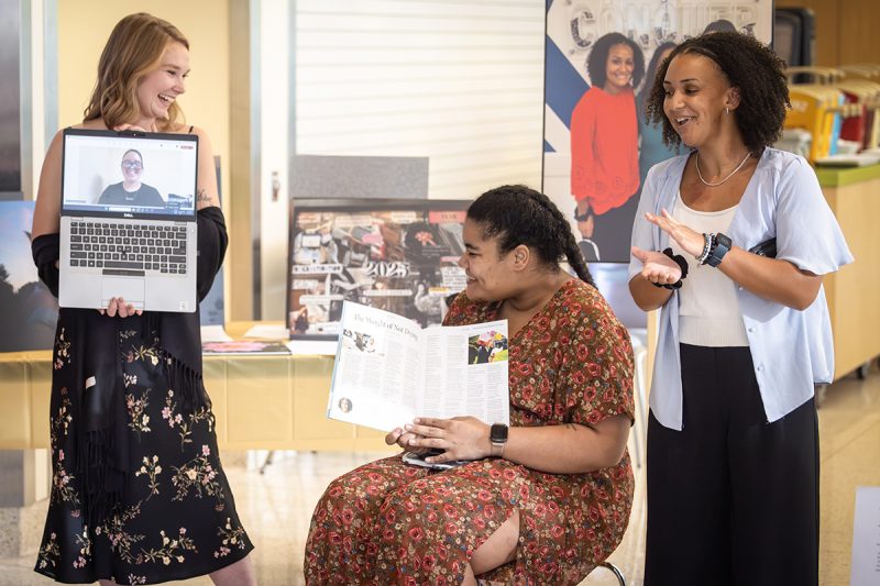 Three women smile and interact during a presentation. One woman stands on the left holding a laptop showing a person on a video call. Another woman sits in the center holding open a magazine article. The third woman stands on the right, clapping and smiling. Posters and display boards are visible in the background.