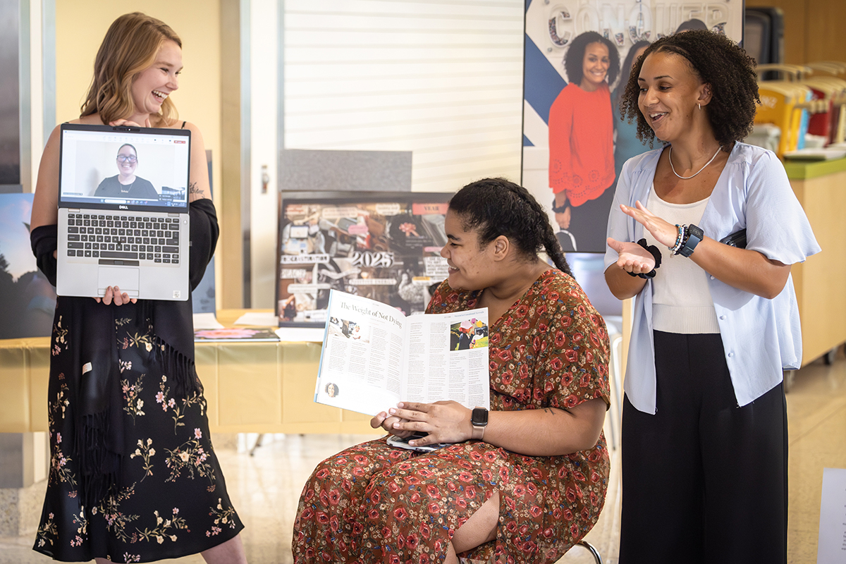 Three women smile and interact during a presentation. One woman stands on the left holding a laptop showing a person on a video call. Another woman sits in the center holding open a magazine article. The third woman stands on the right, clapping and smiling. Posters and display boards are visible in the background.