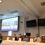 A group of individuals sits below a projector screen as they participate in a discussion panel.