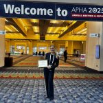 A medical student holds an award certificate and stands below a banner with the words "Welcome to APHA 2025."
