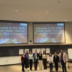 A large group of people stands in the center of a lecture hall below two projector screens.