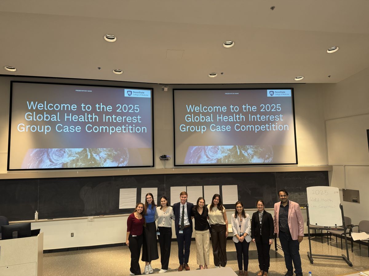 A large group of people stands in the center of a lecture hall below two projector screens.