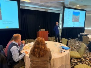 A man stands at the center of a room by a podium and two projector screens.