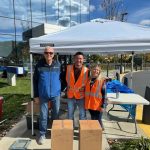Three people wearing orange safety vests stand smiling under a white canopy tent outdoors on a sunny day.