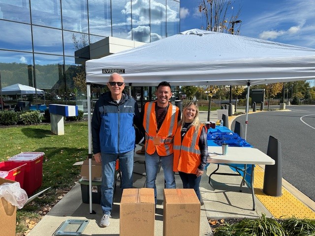 Three people wearing orange safety vests stand smiling under a white canopy tent outdoors on a sunny day.