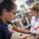 A nurse wearing navy scrubs and glasses draws blood from a patient seated in a medical chair.