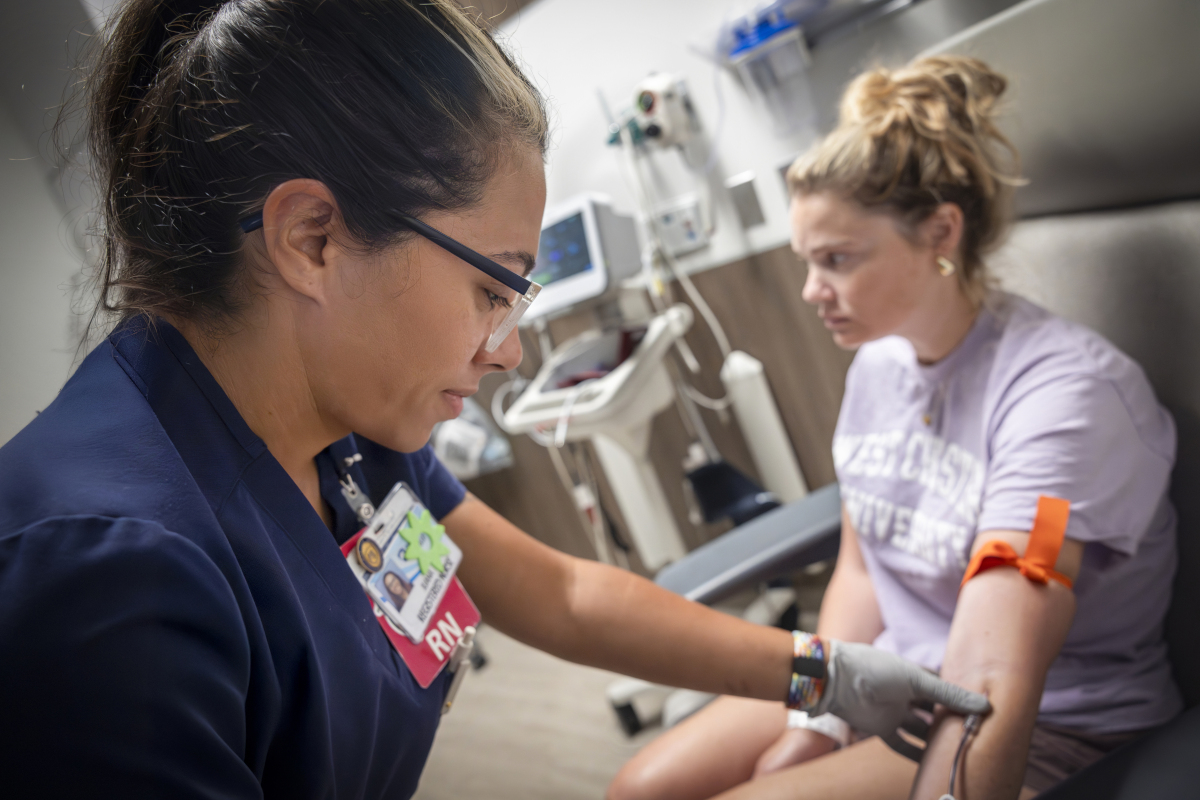 A nurse wearing navy scrubs and glasses draws blood from a patient seated in a medical chair.