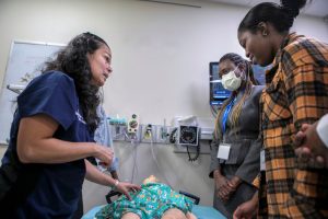 Dr. Lilia Reyes, left touches a mannequin in a simulation lab as she talks to two women. Reyes is wearing scrubs. The woman in the middle is wearing a face mask, and both women are wearing lanyards with nametags. Behind them are a whiteboard, clinical equipment and a monitor.