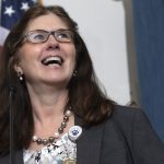 A close-up of Michele Szkolnicki speaking at a lectern. An American flag and curtain are in the background.