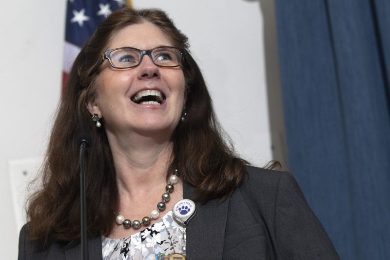 A close-up of Michele Szkolnicki speaking at a lectern. An American flag and curtain are in the background.