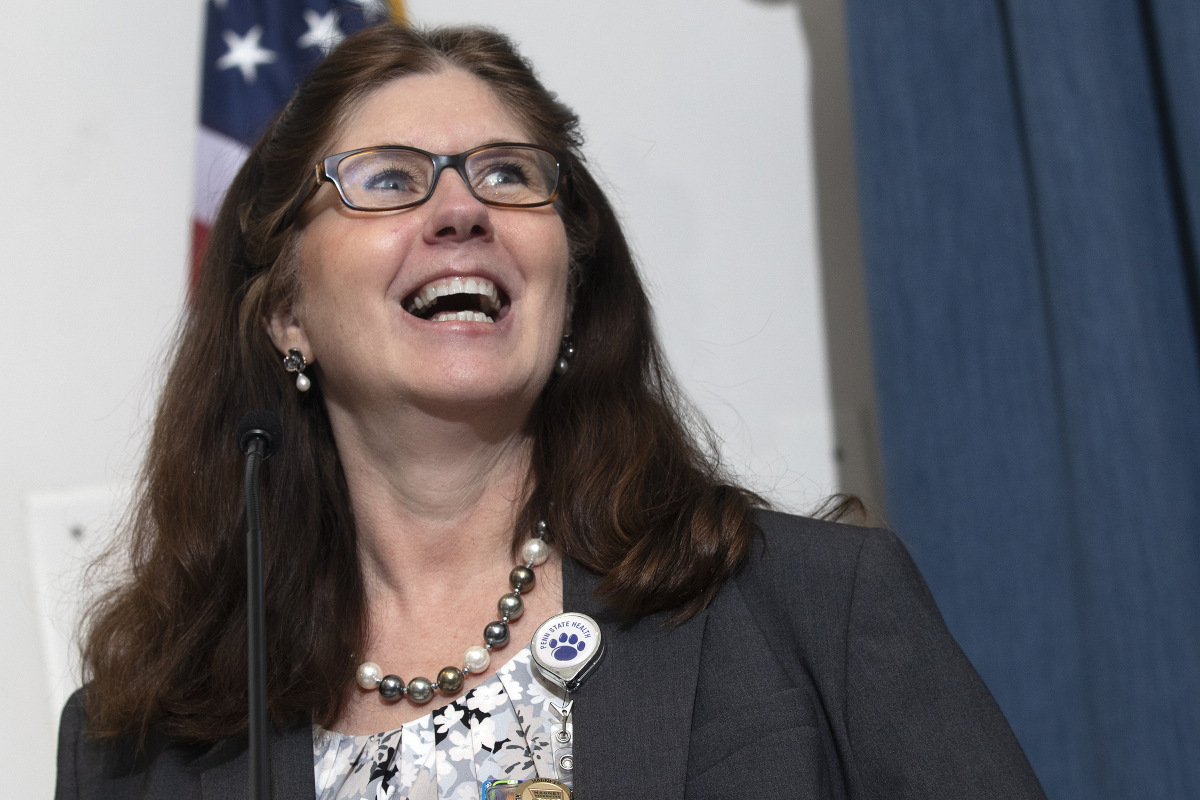 A close-up of Michele Szkolnicki speaking at a lectern. An American flag and curtain are in the background.
