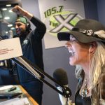 A woman wearing a black hat with feather accents laughs into a microphone while broadcasting in a radio studio. Behind her, a man wearing headphones and a backward cap raises his arms in celebration.