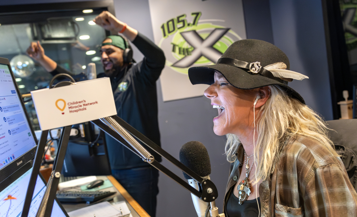 A woman wearing a black hat with feather accents laughs into a microphone while broadcasting in a radio studio. Behind her, a man wearing headphones and a backward cap raises his arms in celebration.
