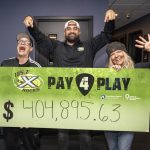 Three smiling radio hosts stand indoors holding a large green “105.7 The X Rocks Pay 4 Play” sign showing the fundraising total of $404,895.63.