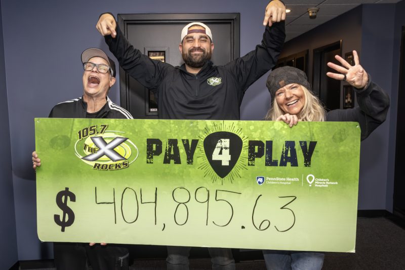 Three smiling radio hosts stand indoors holding a large green “105.7 The X Rocks Pay 4 Play” sign showing the fundraising total of $404,895.63.