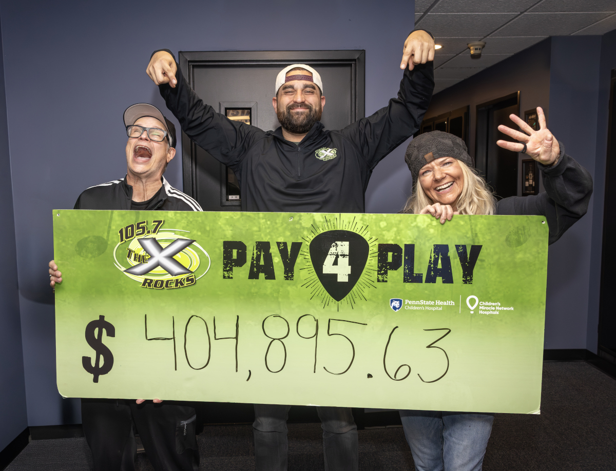 Three smiling radio hosts stand indoors holding a large green “105.7 The X Rocks Pay 4 Play” sign showing the fundraising total of $404,895.63.