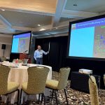 A man stands at the center of a conference room gesturing to a projector screen.