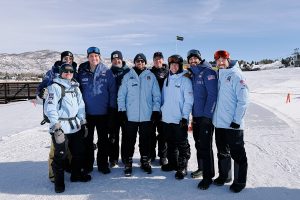 Dr. Neil Blanchard stands in the center of a group of eight other men in ski jackets with U.S. Ski and Snowboard Team and American flag patches on them. They are at a ski resort and wearing hats and ski goggles.