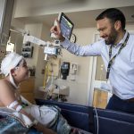 Dr. Brent O'Neill, right, wearing a shirt and tie, raises a finger and smiles at a boy in a hospital bed. The boy is looking up at him. The boy’s head is covered in bandages, he has an IV in his arm and has a blanket on him. Behind them are a monitor and medical equipment.