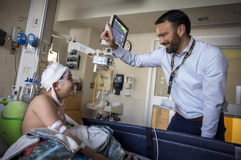 Dr. Brent O'Neill, right, wearing a shirt and tie, raises a finger and smiles at a boy in a hospital bed. The boy is looking up at him. The boy’s head is covered in bandages, he has an IV in his arm and has a blanket on him. Behind them are a monitor and medical equipment.