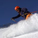 Male skier rides in the deep snow on a sunny day at a ski resort. He is wearing a helmet. A fence is on his left.