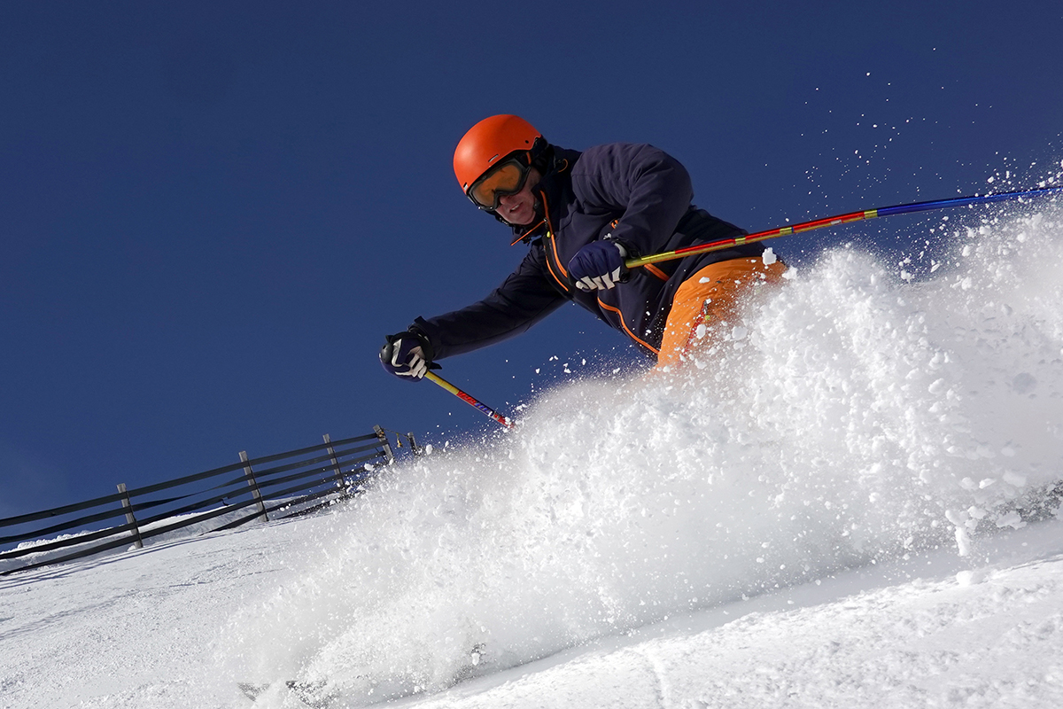 Male skier rides in the deep snow on a sunny day at a ski resort. He is wearing a helmet. A fence is on his left.