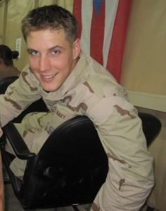 A young man in a U.S. Navy uniform smiles while sitting sideways in a chair inside a military tent. A flag hangs in the background.