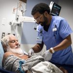 An ED technician tends to a patient laying in a bed. Various medical equipment is mounted to the wall in the background.
