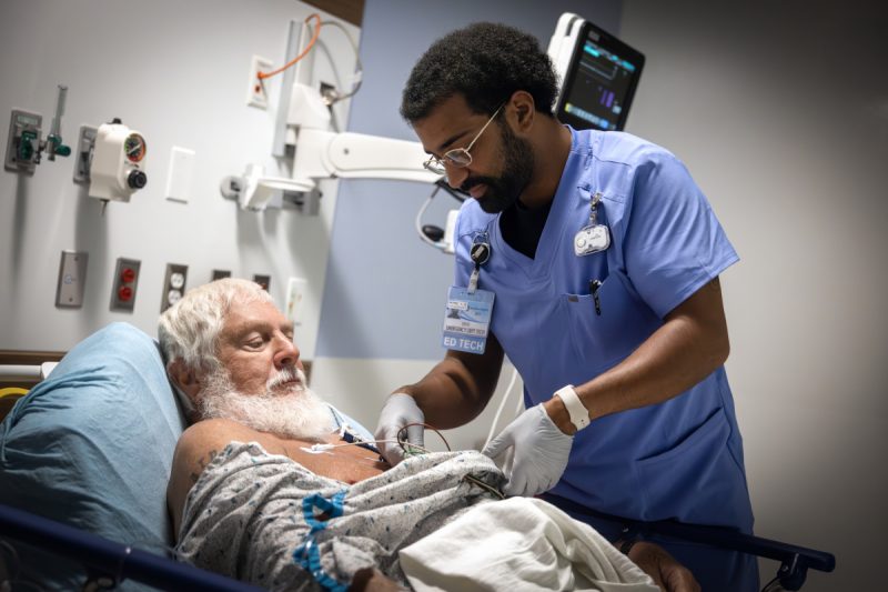An ED technician tends to a patient laying in a bed. Various medical equipment is mounted to the wall in the background.