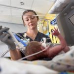 A respiratory therapist wearing scrubs and gloves adjusts breathing tubes for a premature infant lying in an incubator.