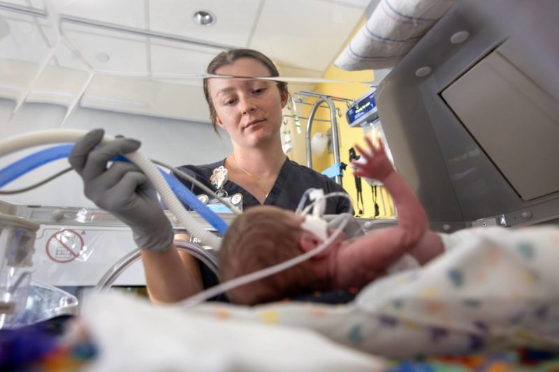A respiratory therapist wearing scrubs and gloves adjusts breathing tubes for a premature infant lying in an incubator.