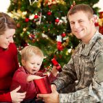 A woman wearing red and a man wearing a U.S. Army uniform sit with their toddler son as he opens a present in front of a Christmas tree.