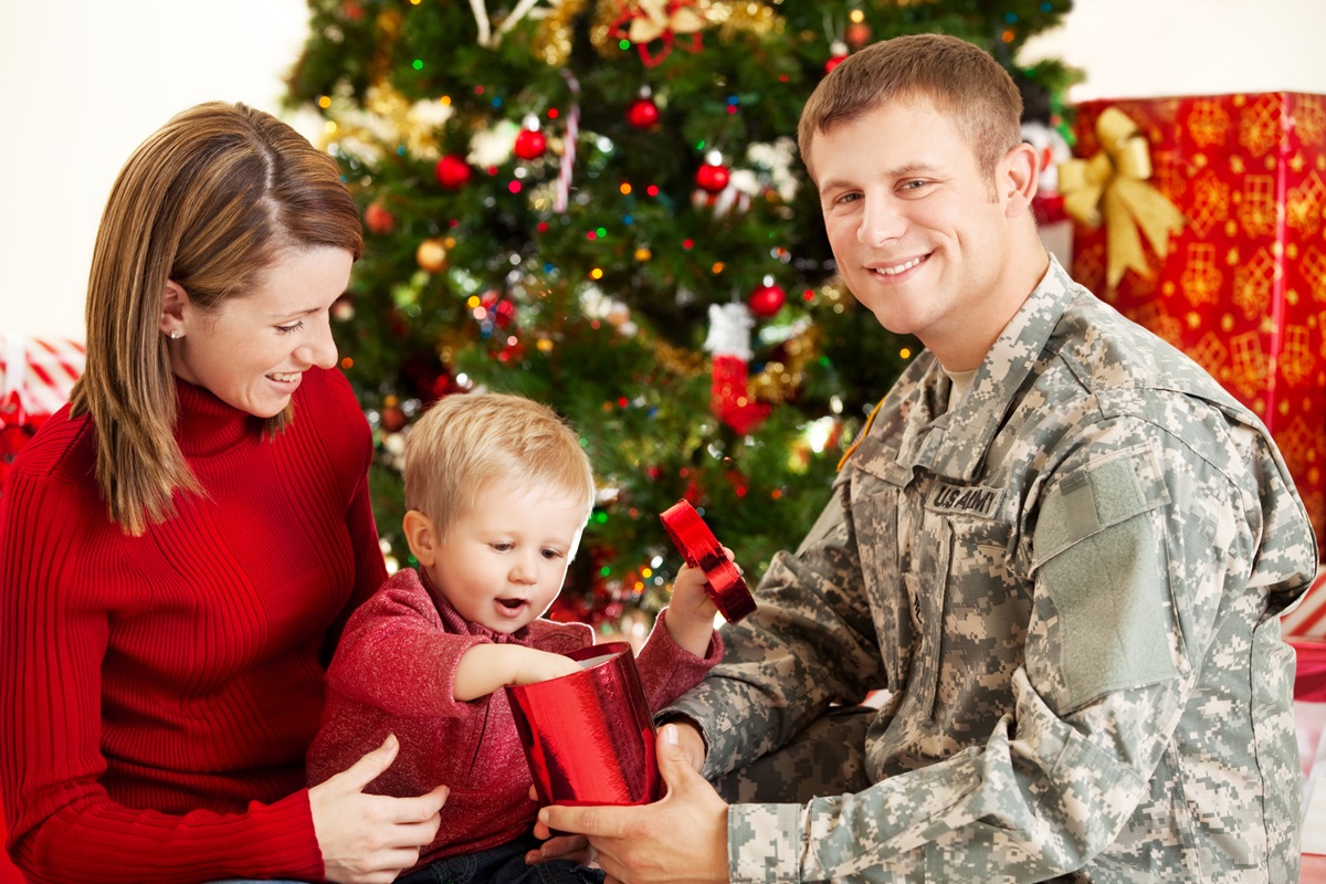 A woman wearing red and a man wearing a U.S. Army uniform sit with their toddler son as he opens a present in front of a Christmas tree.