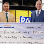 Two men in professional business attire smile as they pose for a photo with an oversized check in the amount of $355,000 made out from Mid Penn Bank to Penn State Health Urology. A TV station set is in the background.