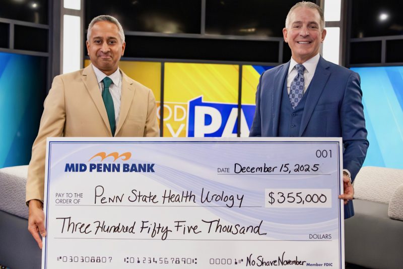 Two men in professional business attire smile as they pose for a photo with an oversized check in the amount of $355,000 made out from Mid Penn Bank to Penn State Health Urology. A TV station set is in the background.