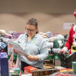 In a room full of gifts such as blankets and books, a woman looks at a children’s book. A man dressed in a seasonal “elf” outfit looks on, as he holds a bag.