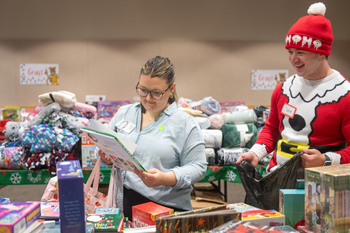 In a room full of gifts such as blankets and books, a woman looks at a children’s book. A man dressed in a seasonal “elf” outfit looks on, as he holds a bag.