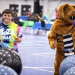 A teen boy and the Nittany Lion mascot use drumsticks to drum on exercise balls in a large gym at Penn State Health RecFest 2025. The boy is wearing a tie-dye T-shirt with the words “3.21 for Life” on it. The Nittany Lion has a scarf.